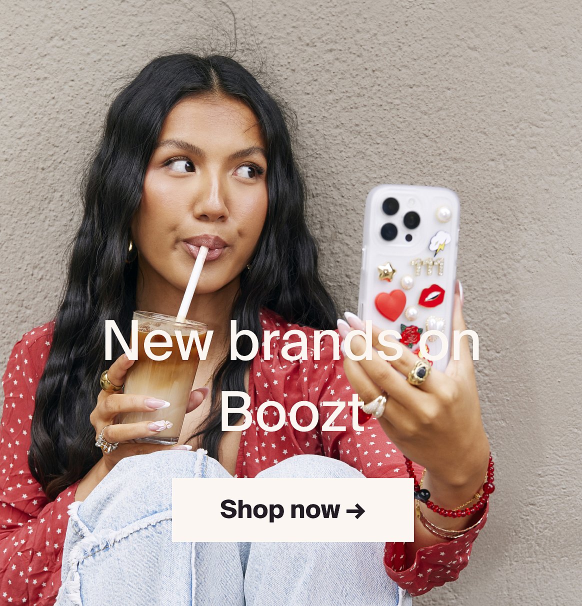 Young woman with long dark hair sips iced coffee, holding a phone with a decorated case, against a gray wall.