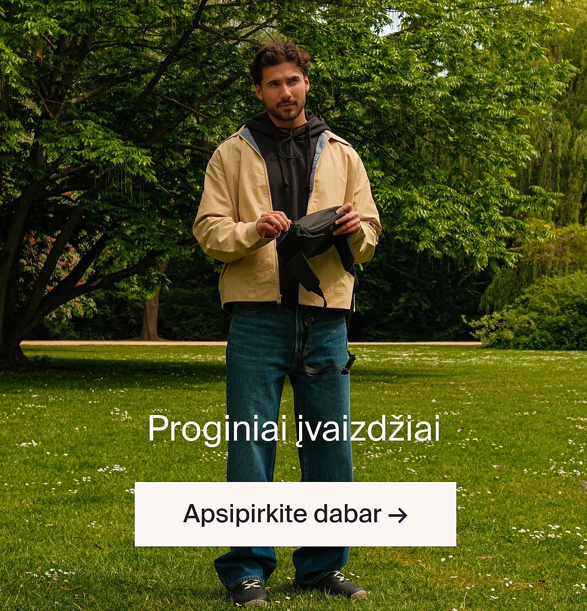 Asmuo su baltais ažūriniais marškinėliais trumpomis rankovėmis ir mėlynais džinsais, rankomis kišenėse, stovi prie šviesios medinės sienos.