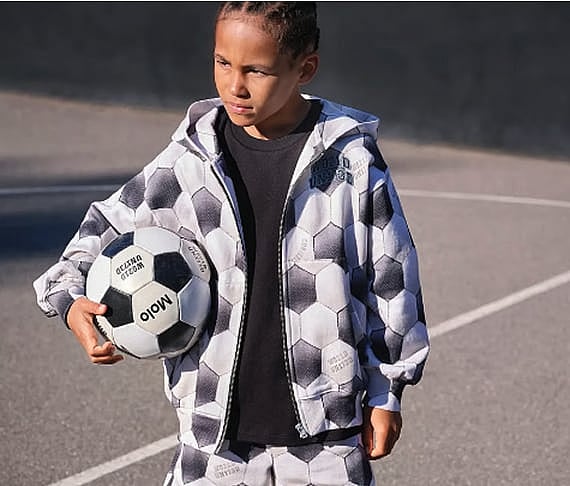 Child with braided hair in a black and white soccer ball hoodie, holding a soccer ball on an outdoor court.