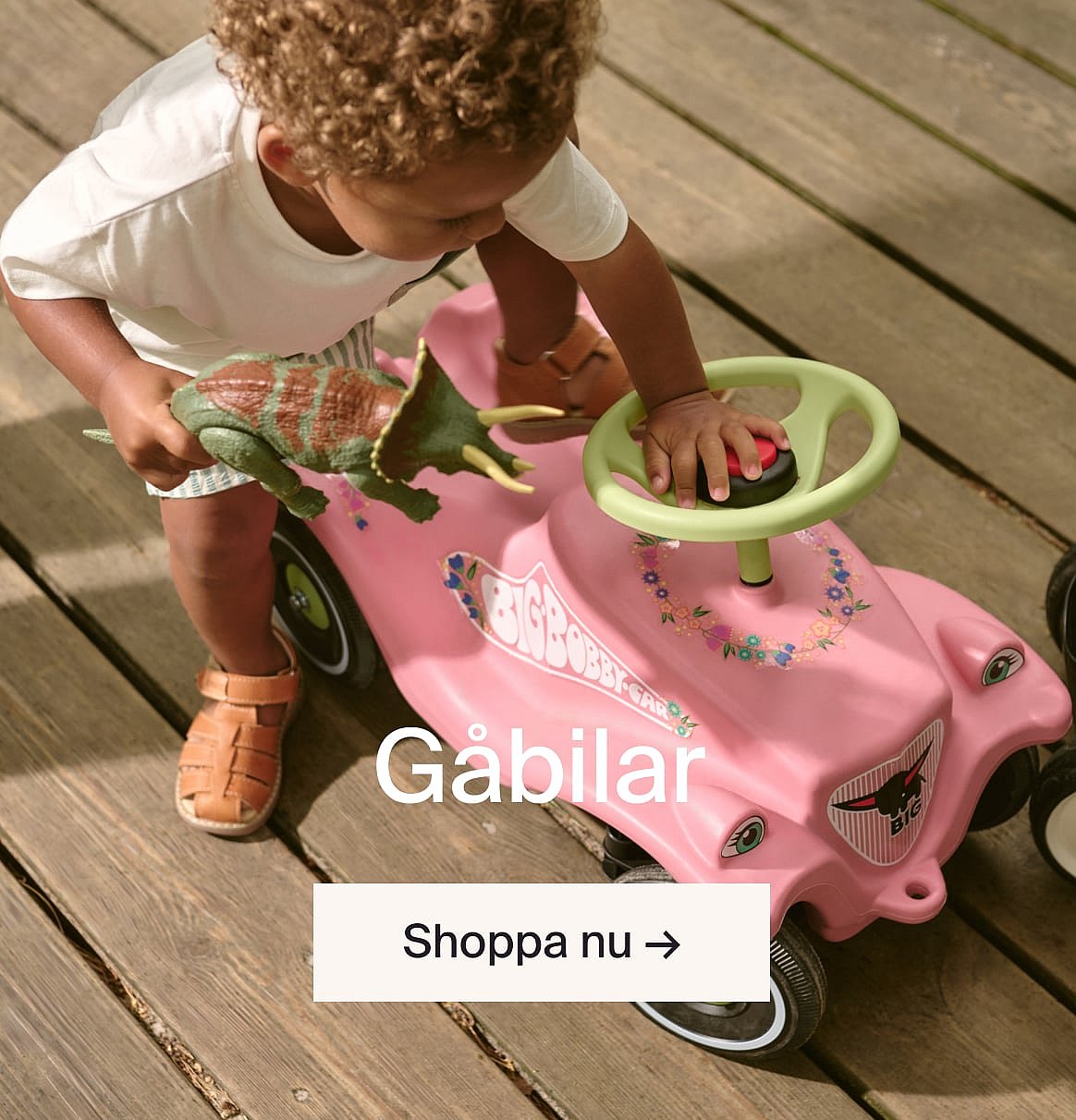 Toddler with curly hair playing with a pink ride-on car and a dinosaur on a wooden deck.