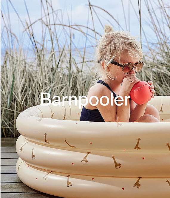 Blonde child in sunglasses sits in a giraffe-patterned inflatable pool, holding a red ball to their mouth, outdoors.