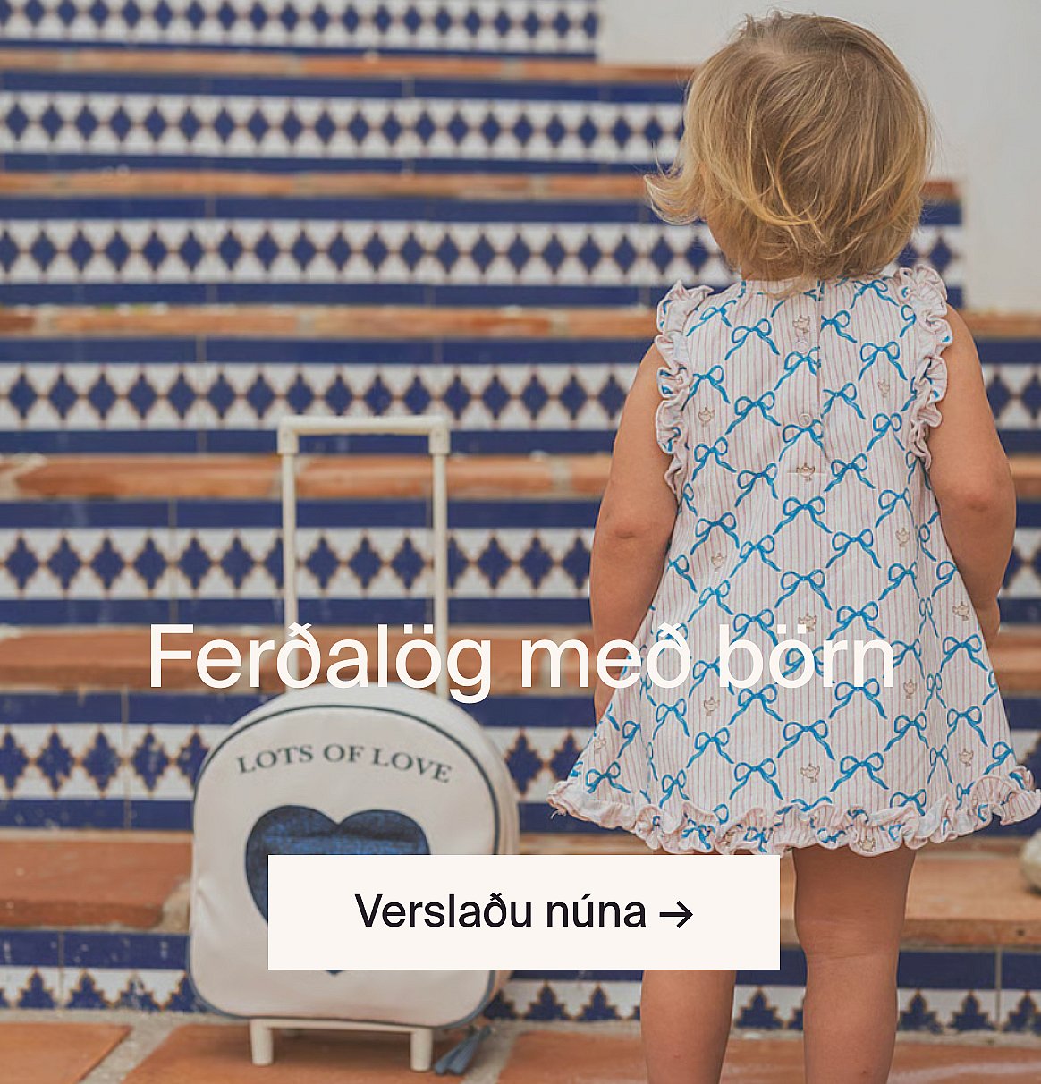 Toddler in a patterned dress stands on tiled steps next to a small suitcase.