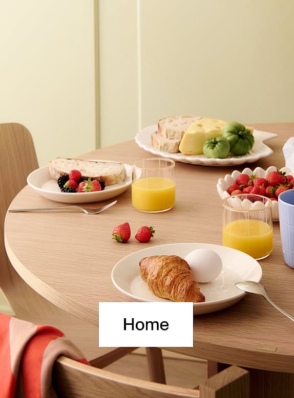 Modern light wood chair and round table with an open book and a light blue coffee mug, against a yellow wall and wood floor.