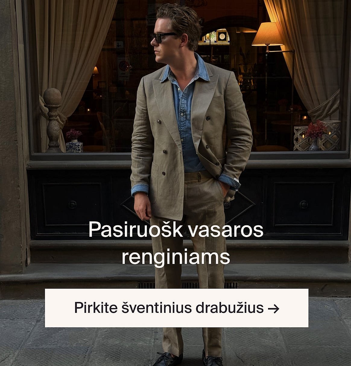 Asmuo su baltais ažūriniais marškinėliais trumpomis rankovėmis ir mėlynais džinsais, rankomis kišenėse, stovi prie šviesios medinės sienos.