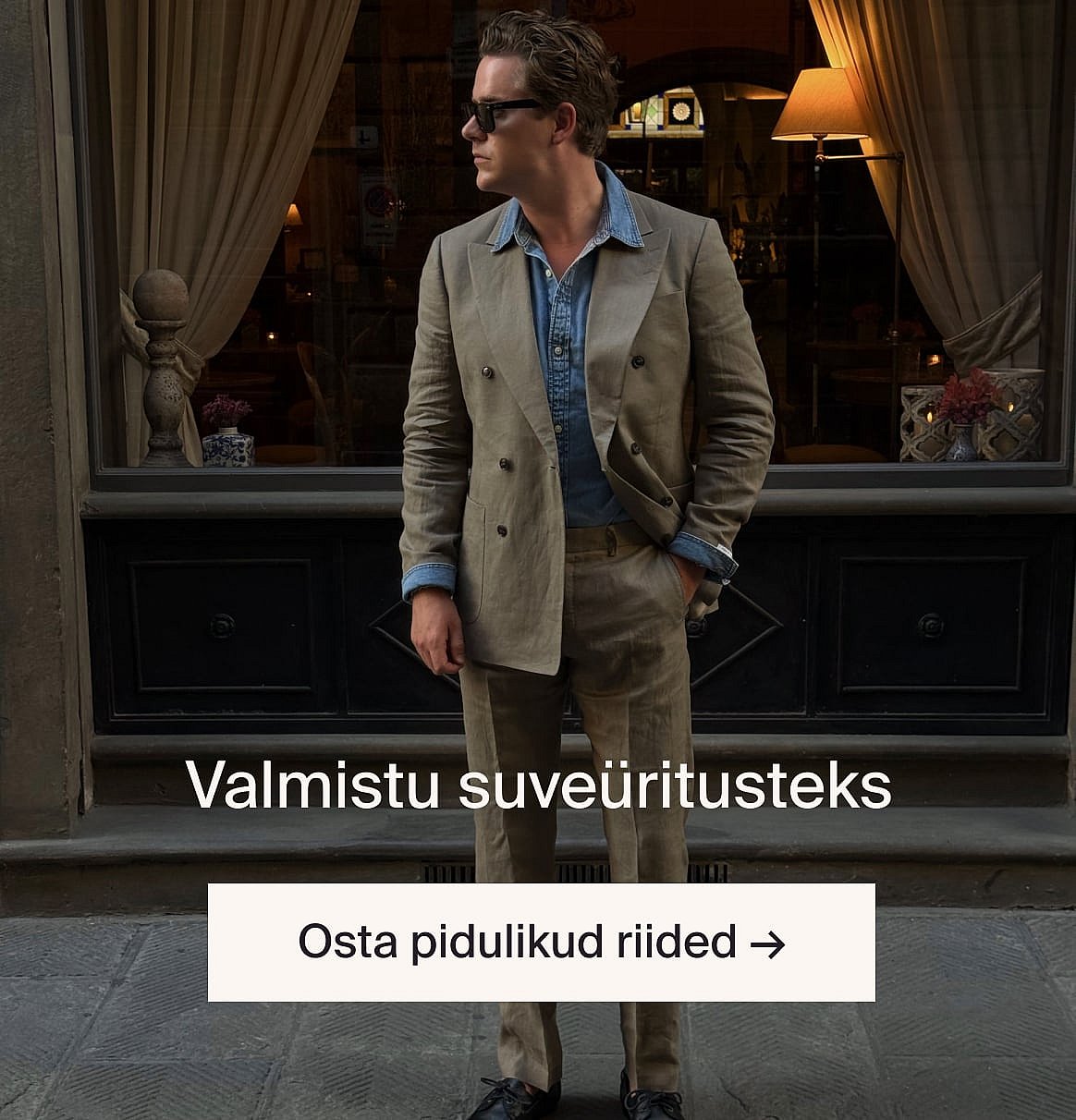 Valges lühikeste varrukatega broderiisärgis ja sinistel teksapükstes, käed taskutes, seistes heleda puitseina vastu.