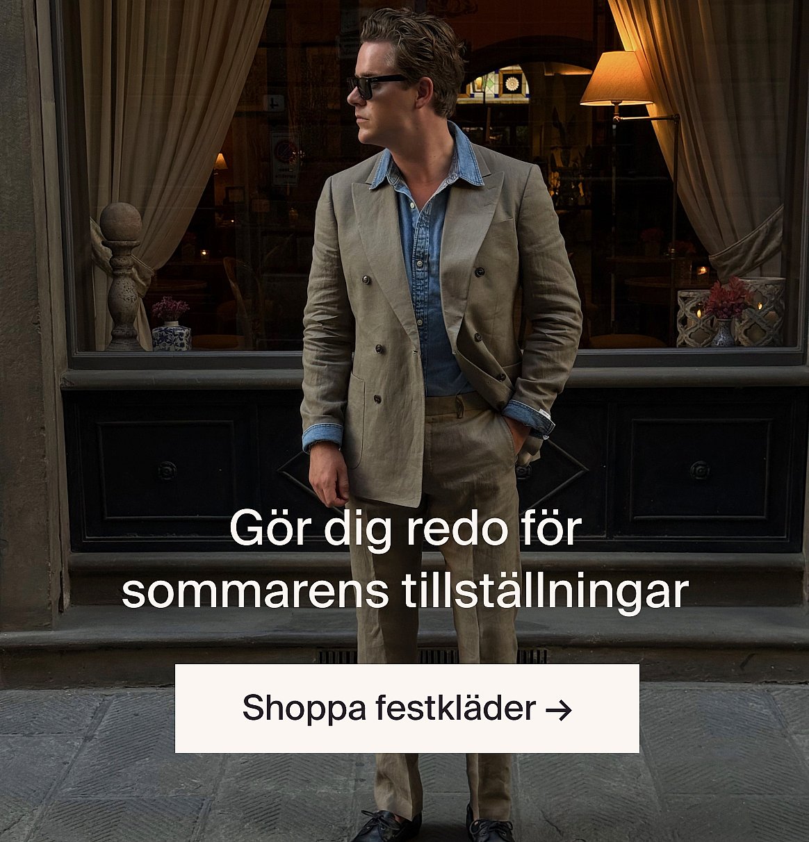 Person i vit kortärmad broderie anglaise-skjorta och blå jeans, händerna i fickorna, står mot en ljus trävägg.