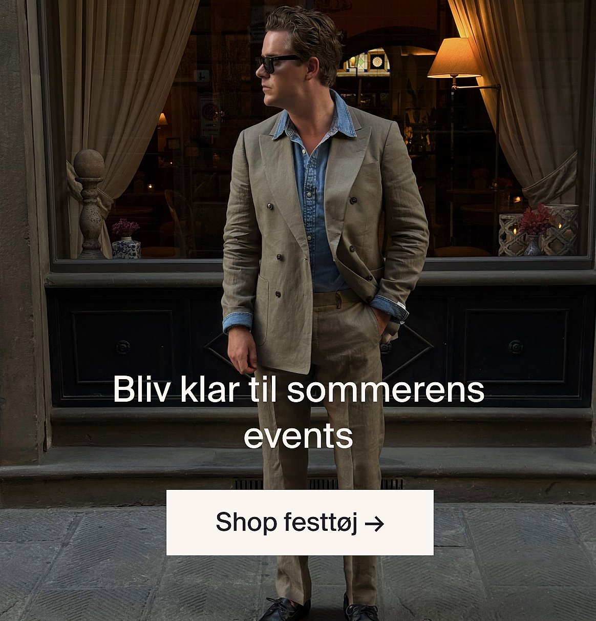 Hvid kortærmet skjorte med broderie anglaise og blå jeans, hænderne i lommerne, stående op ad en lys trævæg.