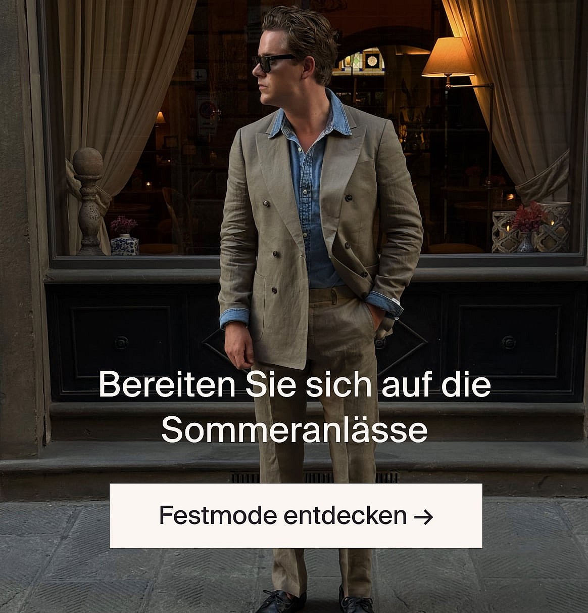 Person in weißem kurzärmeligem Oberteil mit Lochstickerei und blauen Jeans, die Hände in den Hosentaschen, steht vor einer hellen Holzwand.