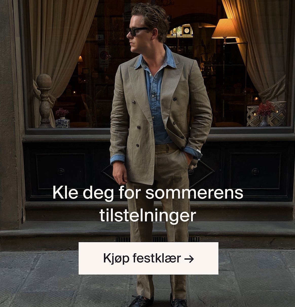 Person i hvit kortermet skjorte med hullbroderi og blå jeans, hender i lommene, står mot en lys trevegg.