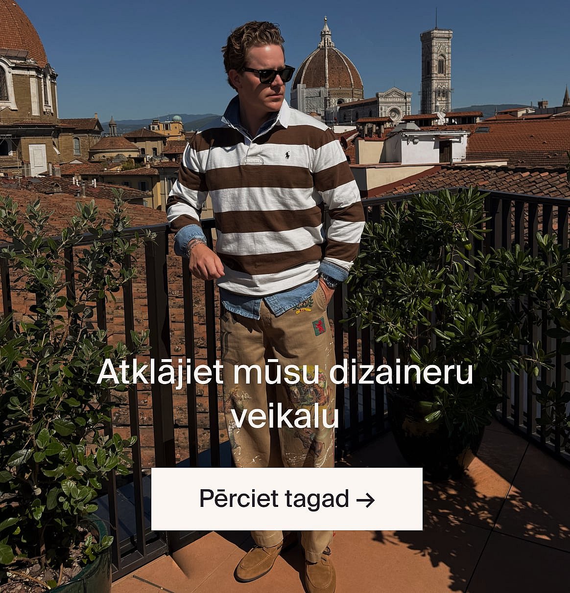 Vīrietis gaišbēšā uzvalkā un zilā kreklā ārā, meža laukumā, pie galda ar zaļumiem.