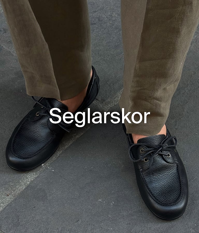En person i bruna shorts, vit skjorta och blå jacka håller i gröna växter utomhus vid en trävägg.