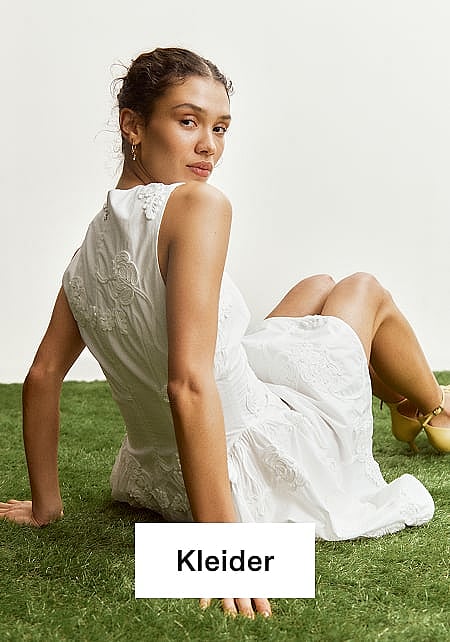 Eine Frau in einem weißen, bestickten, ärmellosen Kleid und gelben Schuhen sitzt auf grünem Gras und schaut zurück. Text: Kleider.