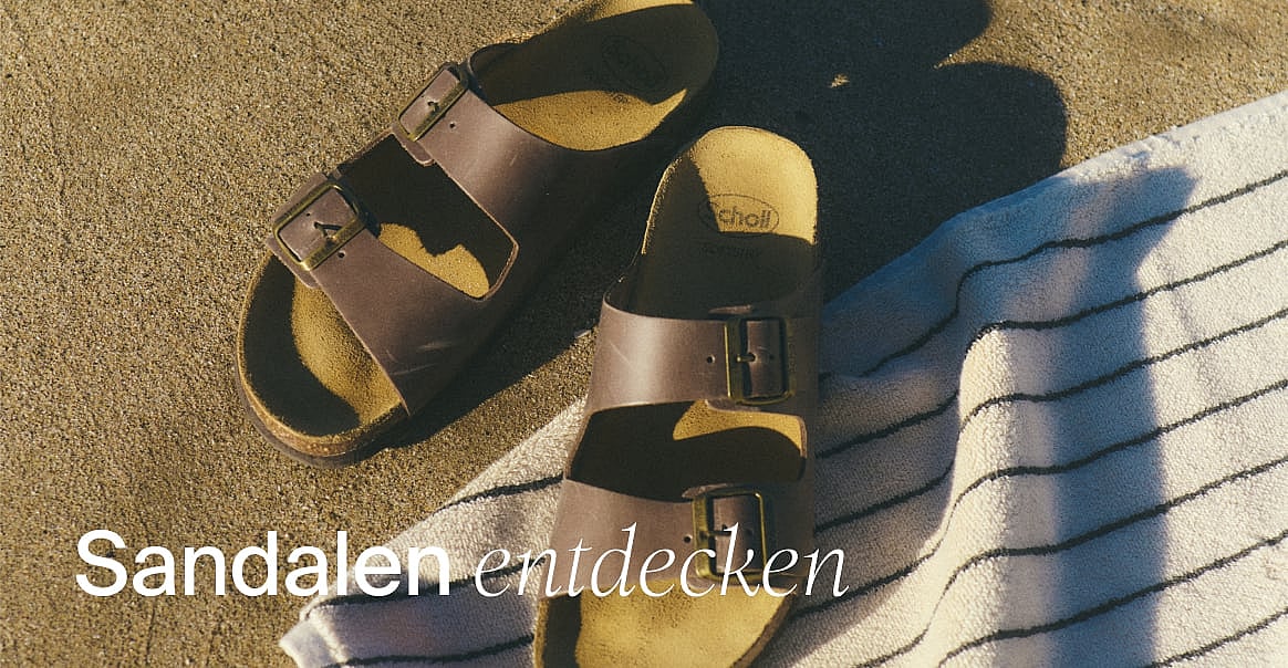 Schwarzer Rollkoffer auf einem gemusterten Boden, neben dem Bein einer Person. Text: „Packe wie ein Profi. Entdecke die Reisekollektion.“
