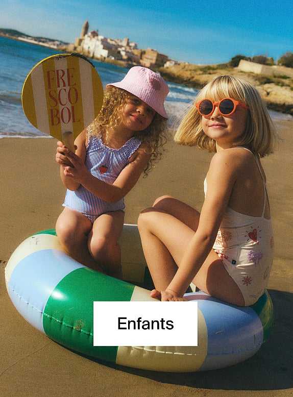 Un jeune garçon en ensemble en jean bleu est assis en tailleur en extérieur, les yeux plissés et souriant.