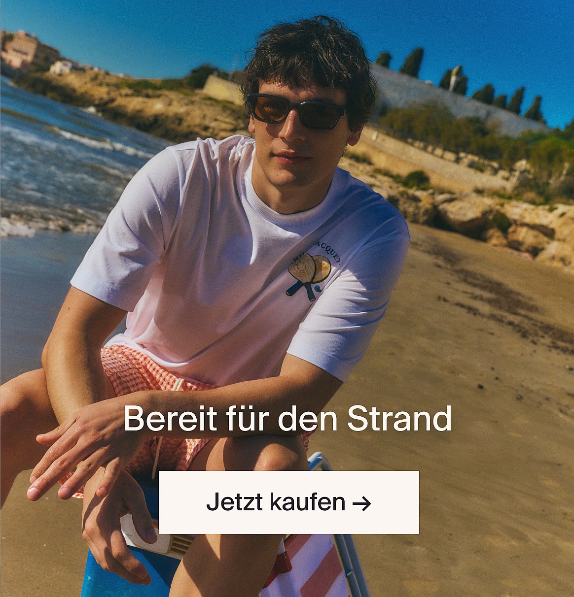 Person in weißem kurzärmeligem Oberteil mit Broderie Anglaise und blauer Jeans, Hände in den Taschen, vor einer hellen Holzwand stehend.