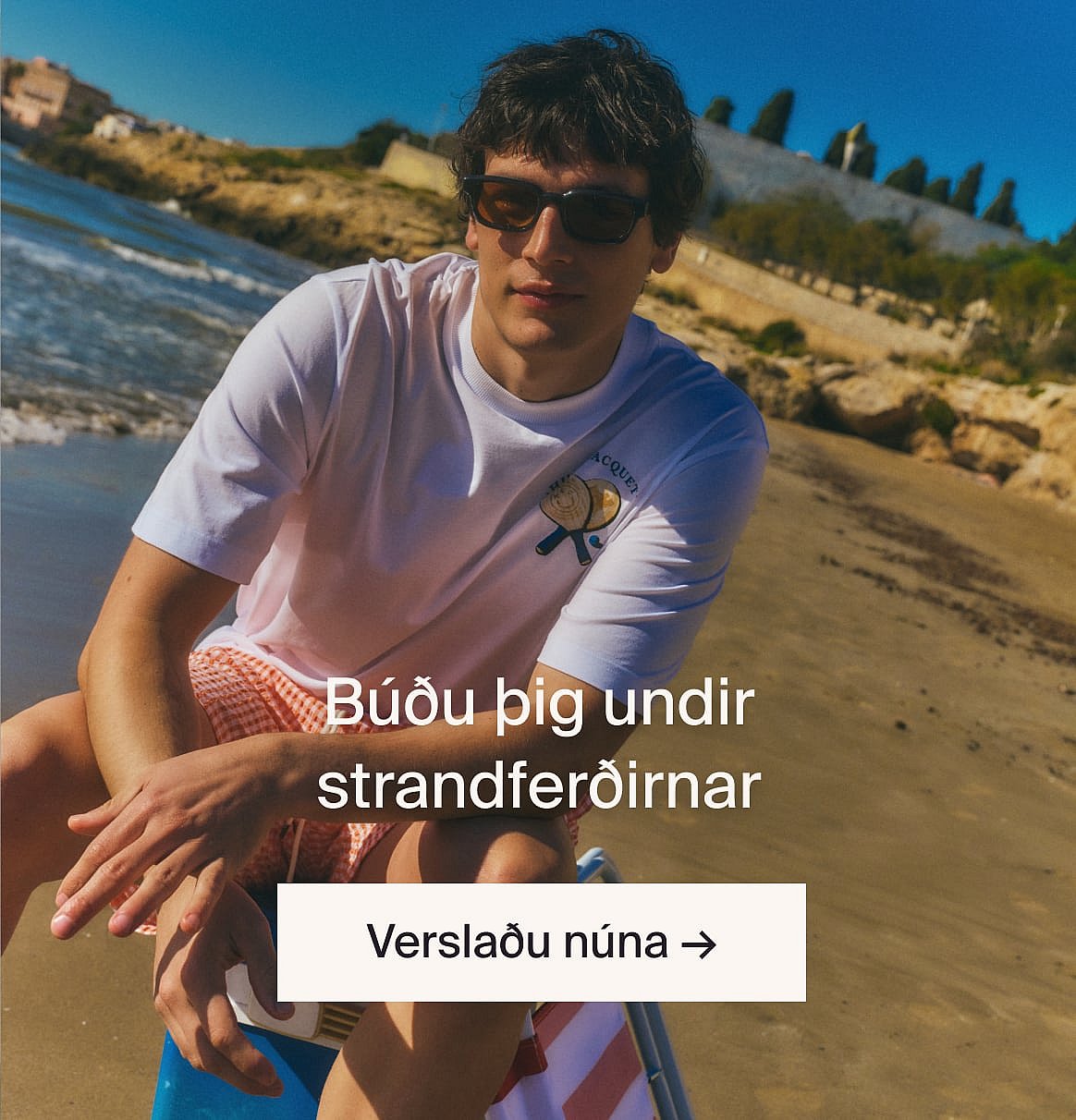 Manneskja í hvítum götóttum stuttermabol og bláum gallabuxum, með hendur í vösum, stendur við ljósan viðarvegg.