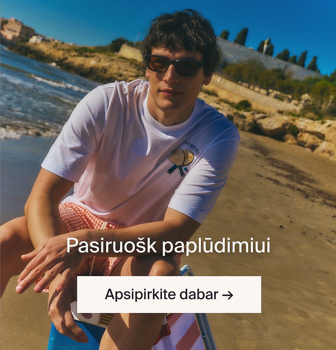Asmuo su baltais ažūriniais marškinėliais trumpomis rankovėmis ir mėlynais džinsais, rankomis kišenėse, stovi prie šviesios medinės sienos.