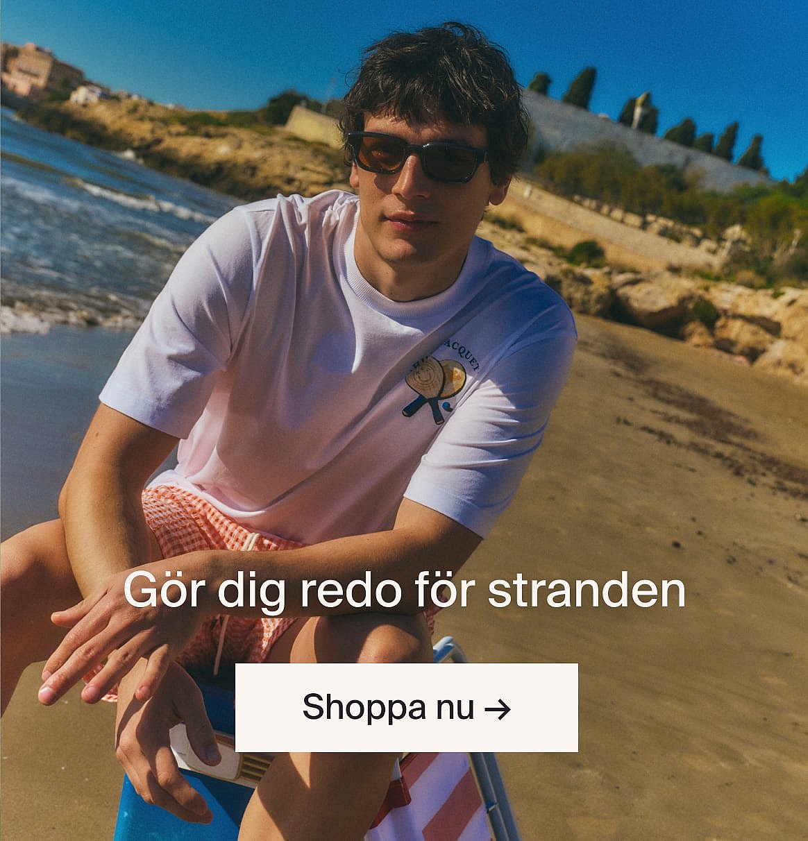 Person i vit kortärmad broderie anglaise-skjorta och blå jeans, händerna i fickorna, står mot en ljus trävägg.