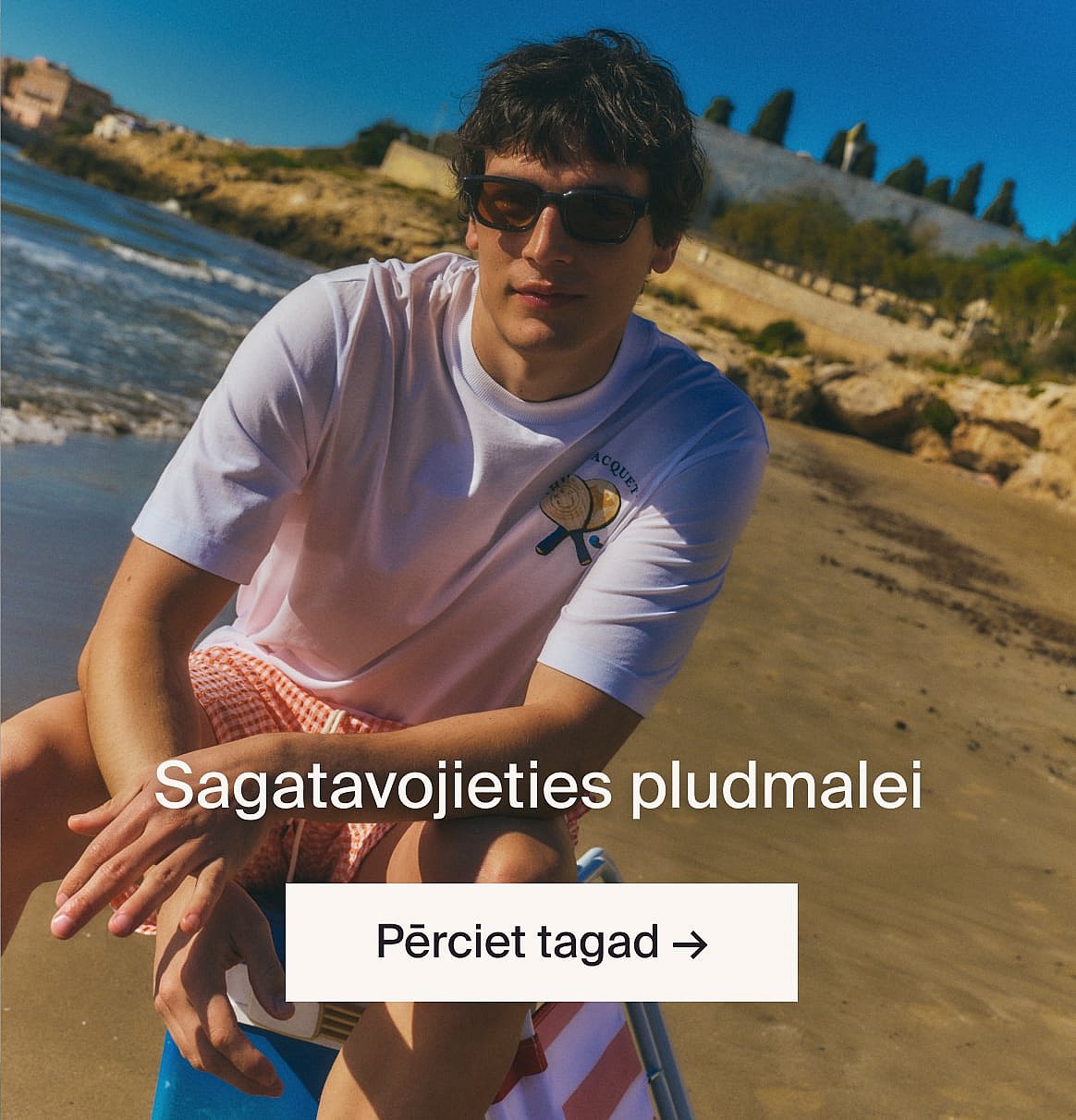 Persona baltā ažūra blūzē ar īsām piedurknēm un zilos džinsos, rokas kabatās, stāv pret gaišu koka sienu.