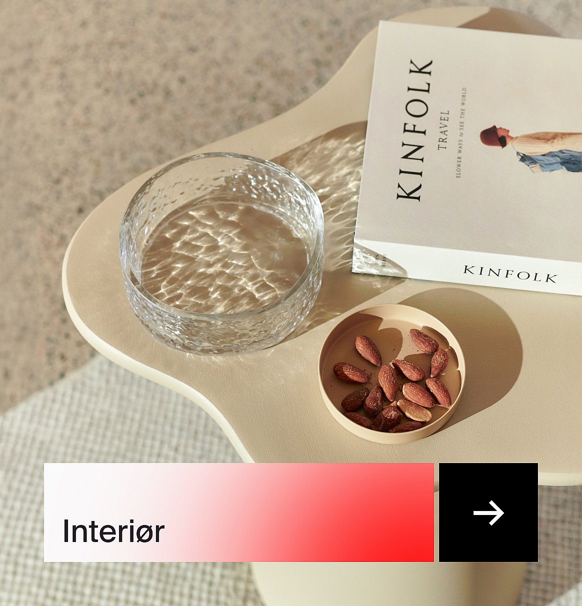 Et beige bord med en skål av strukturert glass, boken «Kinfolk Travel» og mandler, badet i sollys.