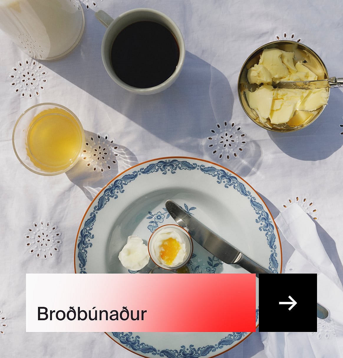 Morgunverður að ofan: kaffi, safi, mjúksoðið egg, smjör og mynstrað fat á hvítum saumuðum borðdúk.