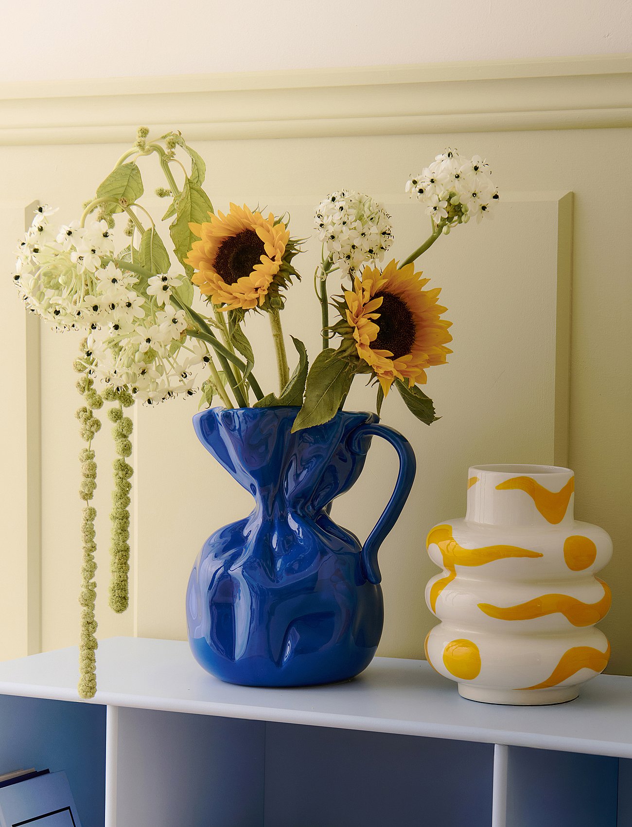 Blue crumpled vase with sunflowers and white flowers, next to a white and yellow patterned vase, on a light blue shelf.