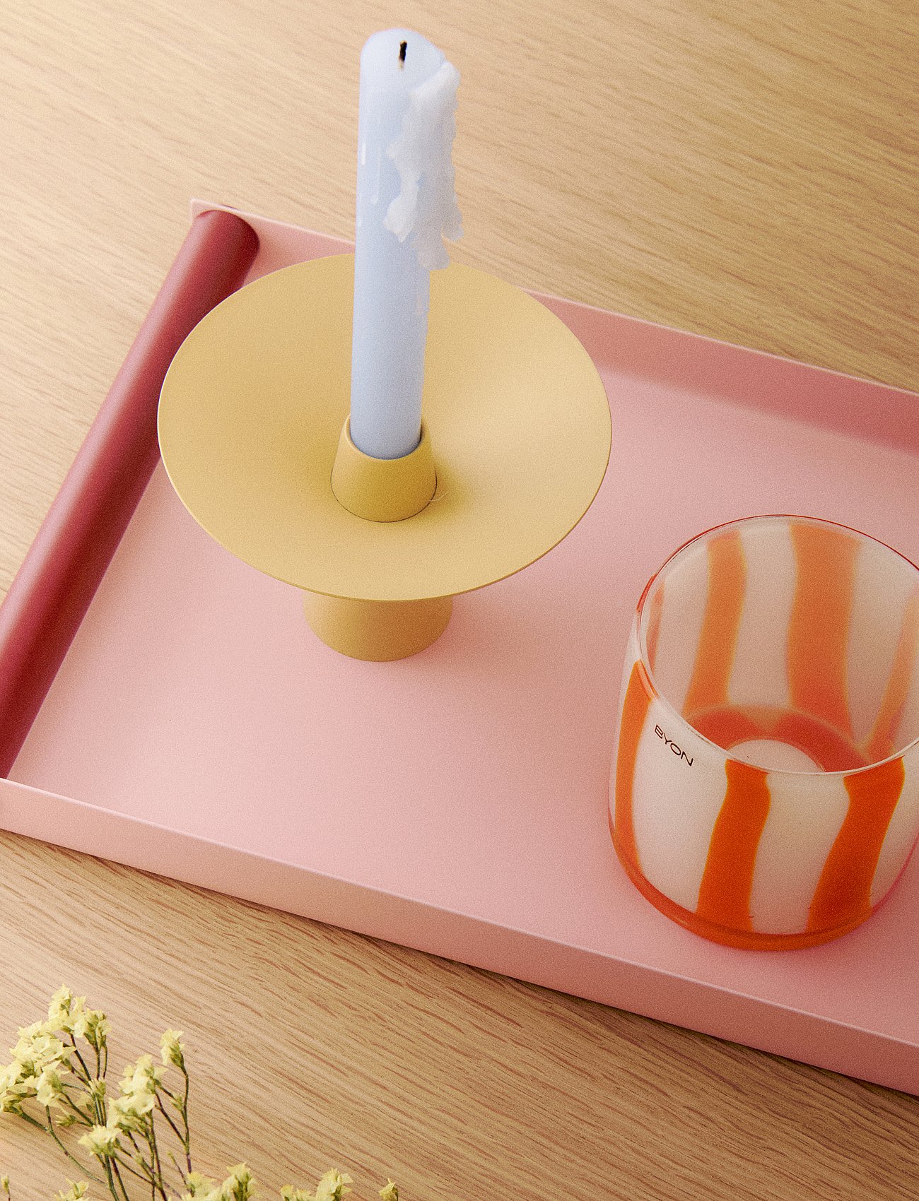 Light blue candle in yellow holder, striped glass, and red cylinder on pink tray, on wood with dried flowers.