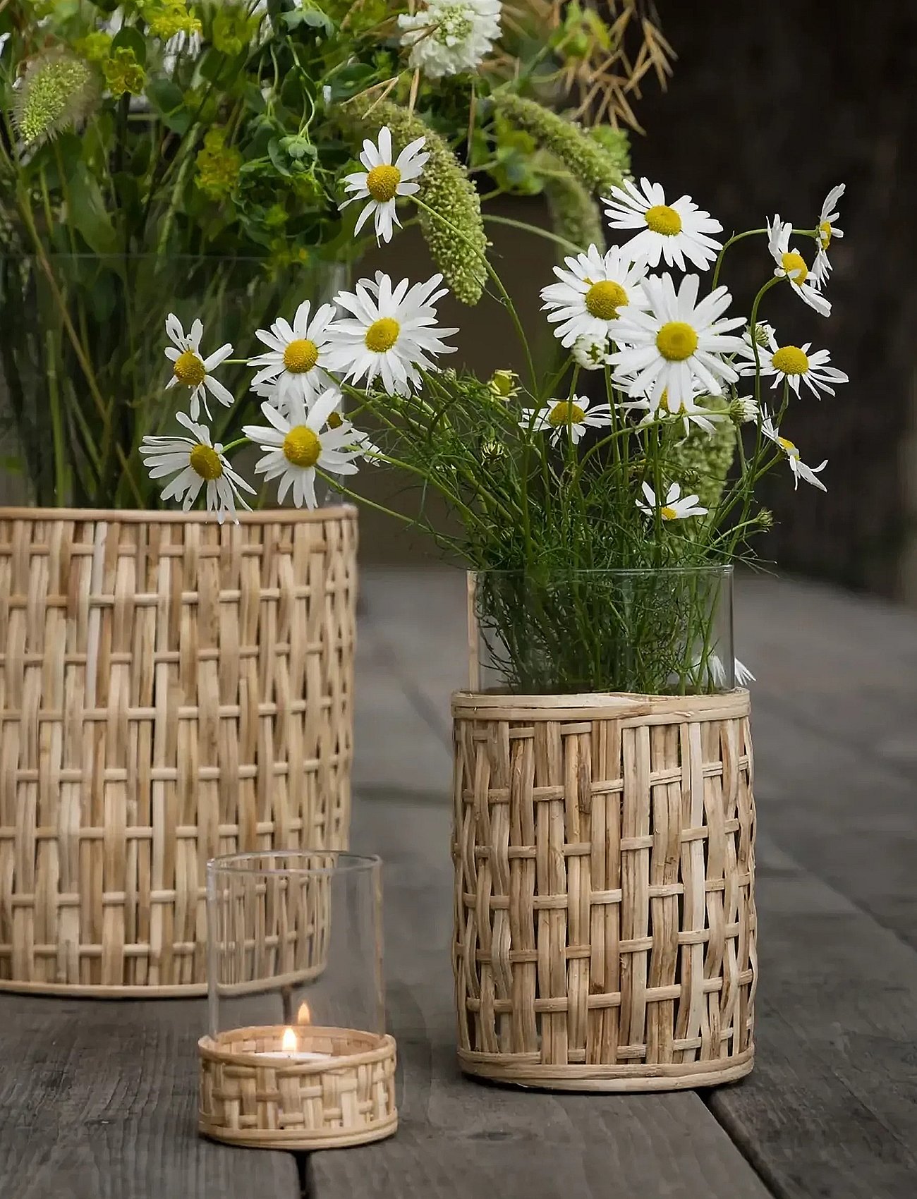 Two woven bamboo containers with white daisies and greenery, and a lit candle in a matching holder, on a wooden surface.