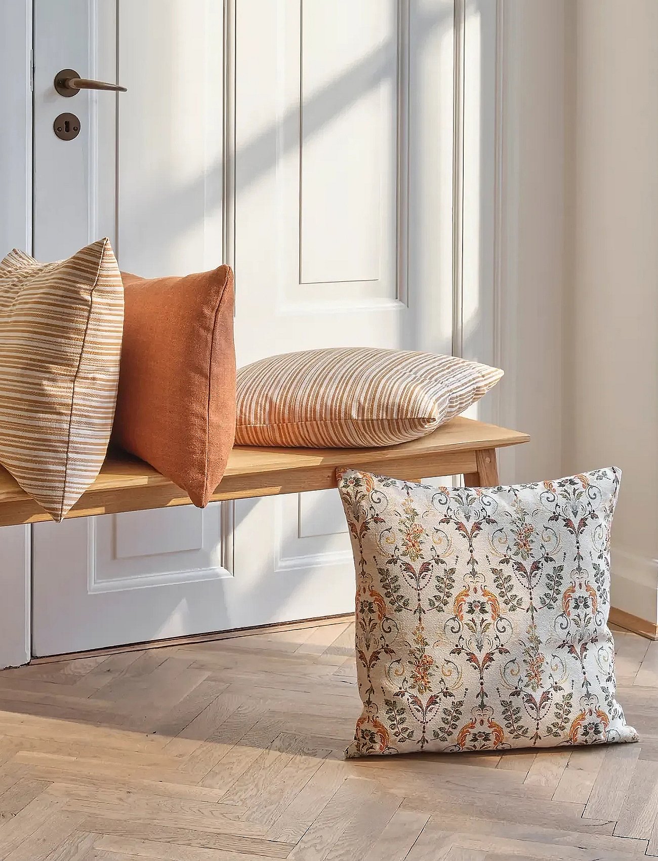 Assorted cushions on a wooden bench and floor, bathed in sunlight, against a white paneled door.