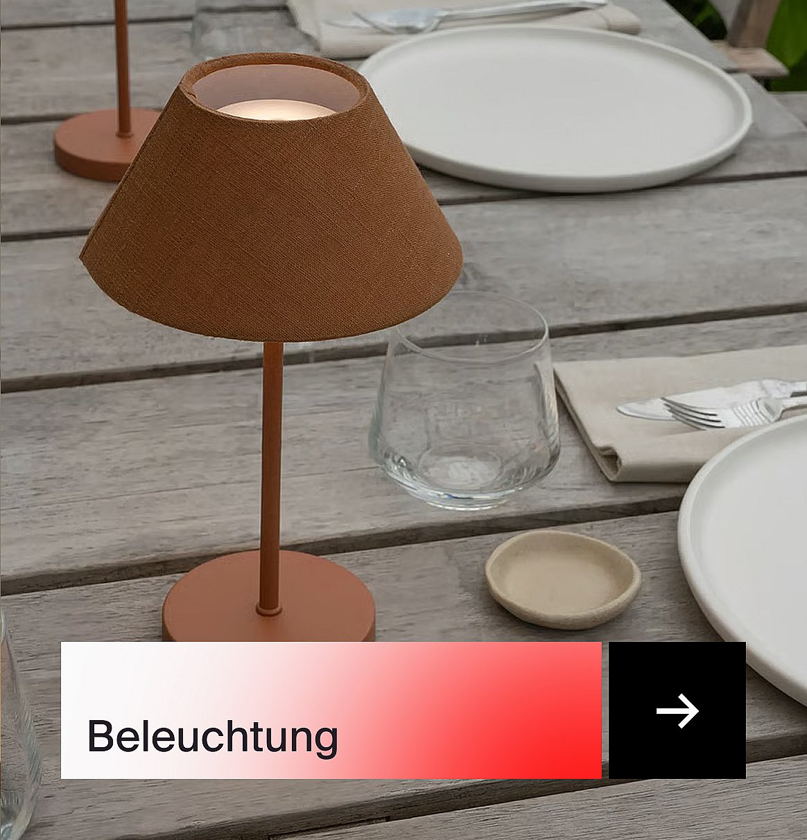 Eine braune Tischlampe leuchtet auf einem hölzernen Esstisch im Freien, der mit weißen Tellern, Gläsern und Besteck gedeckt ist.