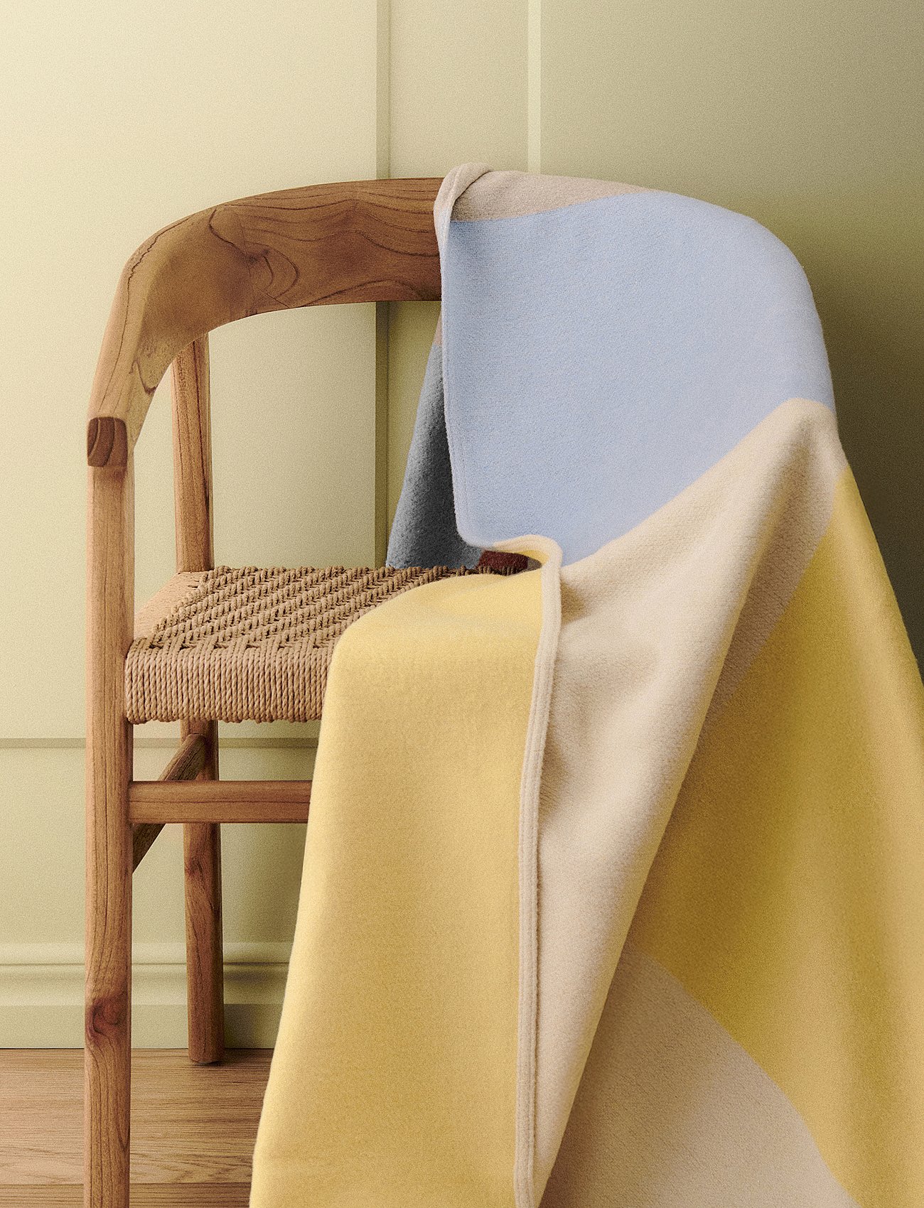 Über einem Holzstuhl mit geflochtener Sitzfläche liegt eine Decke in den Farben Blau, Gelb und Beige, vor einem hellgrünen Hintergrund