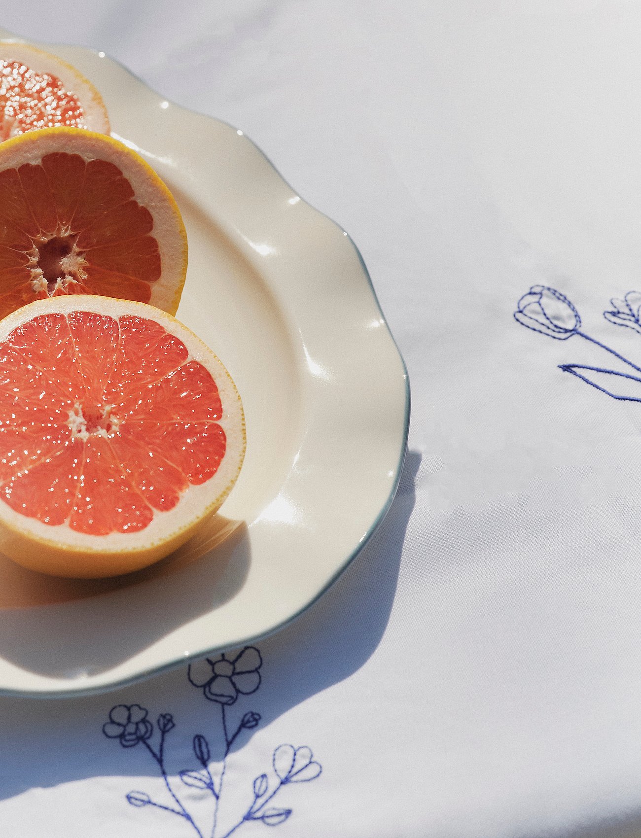 Drei halbierte Grapefruits auf einem gewellten cremefarbenen Teller, auf einer weißen Tischdecke mit blau gestickten Blumen, im Sonnenlicht.