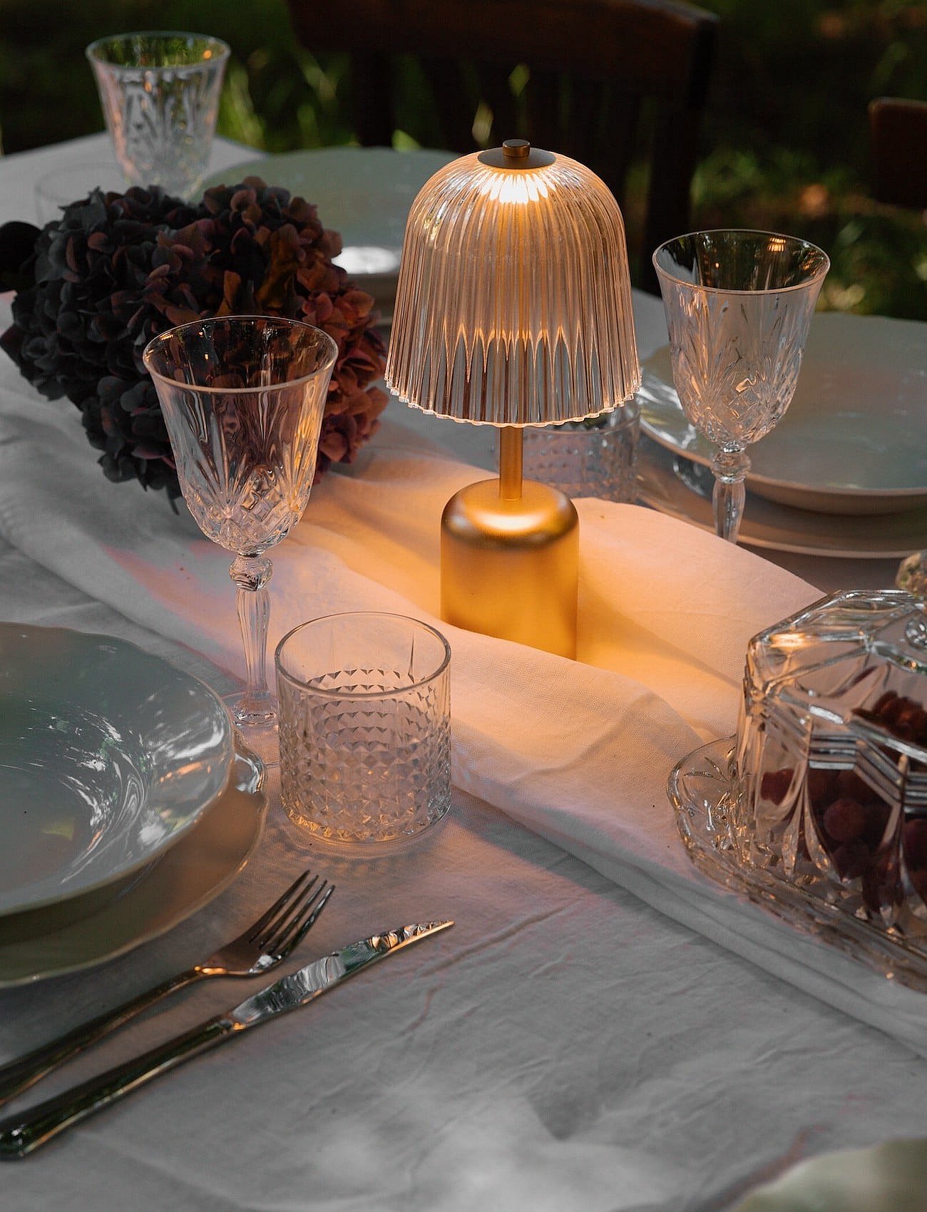 Ein leuchtendes, geripptes Glas und eine tragbare goldene Tischlampe beleuchten einen Esstisch im Freien, der mit Kristallgläsern und -tellern gedeckt ist.