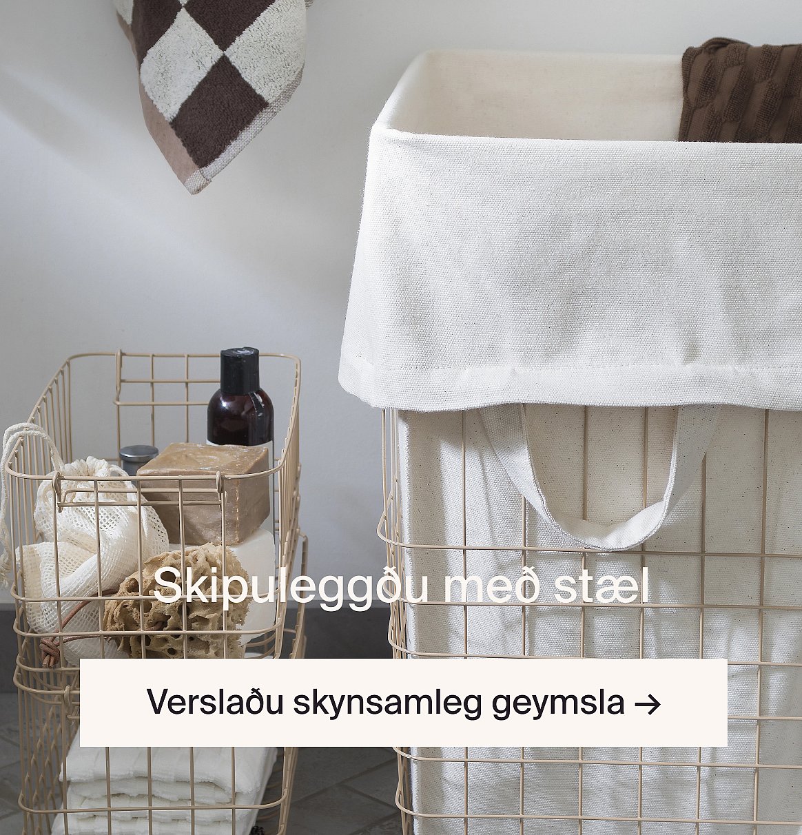 Tvær beigurar geymslukörfur úr vír með hvítum dúkfóðrum. Önnur inniheldur baðvörur, hin er þvottakörfa.
