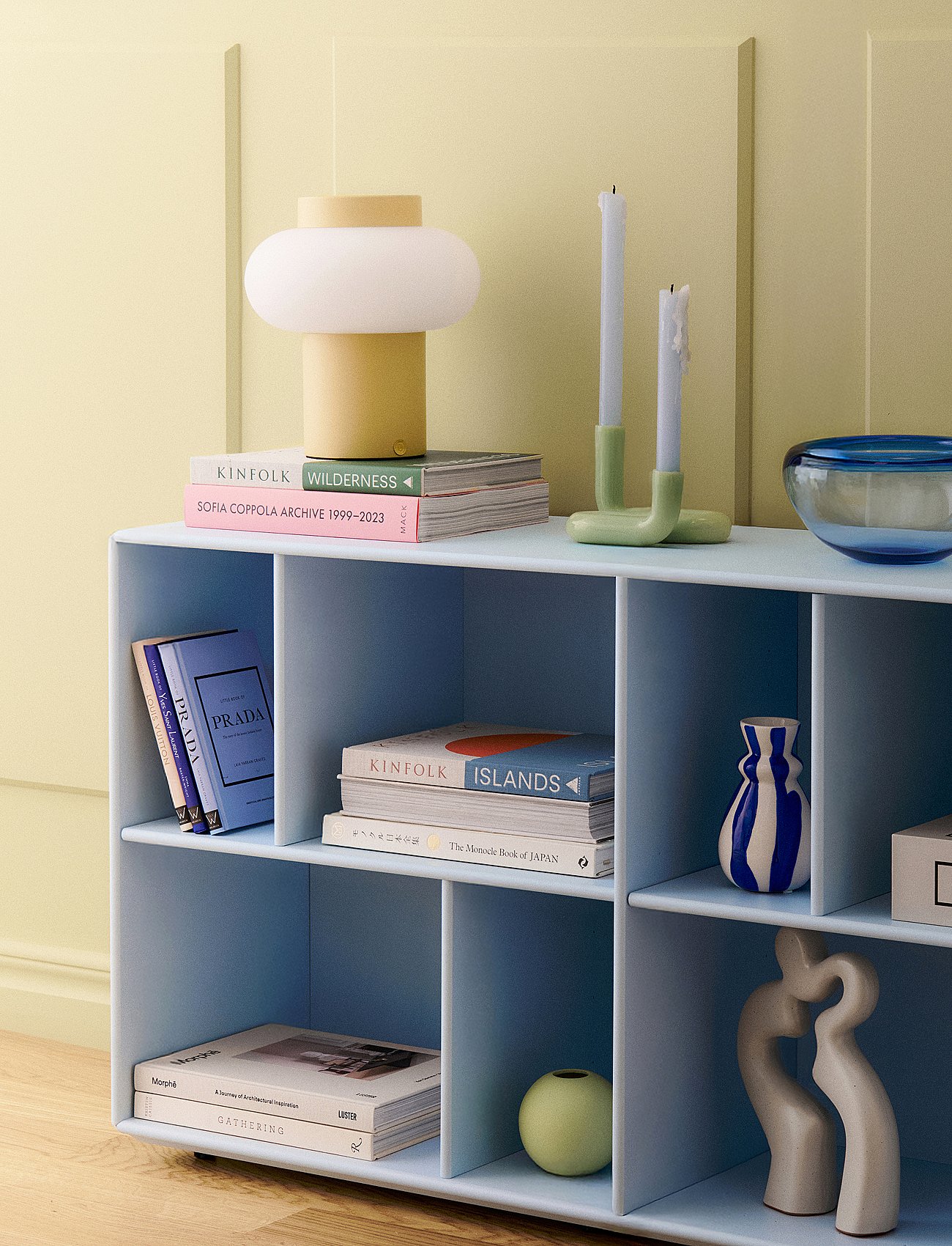 Light blue modular shelving unit against a pale yellow wall, displaying books, a yellow lamp, candles and decoration.
