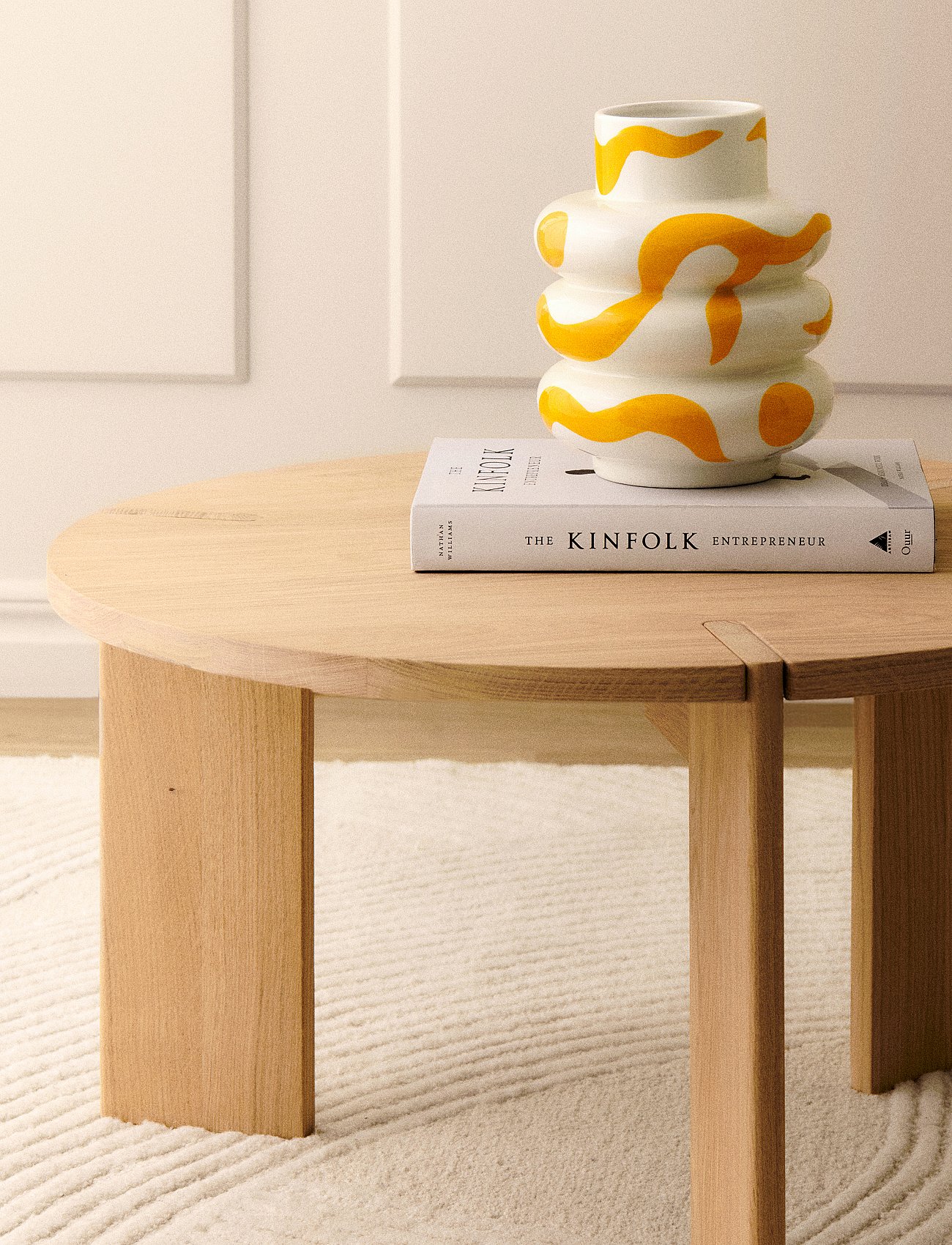 A white vase with yellow patterns and "The Kinfolk Entrepreneur" book on a light wood round table, on a textured rug.