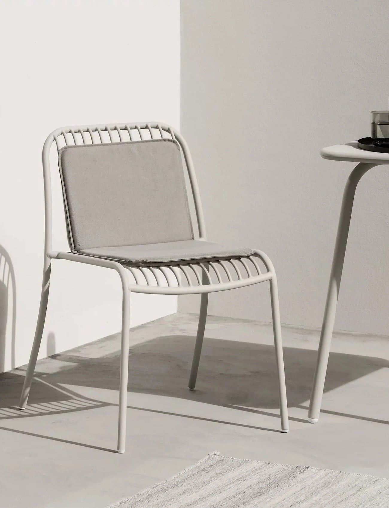 Light gray slatted outdoor metal chair with gray cushions, beside a matching table, on a concrete floor with a rug.