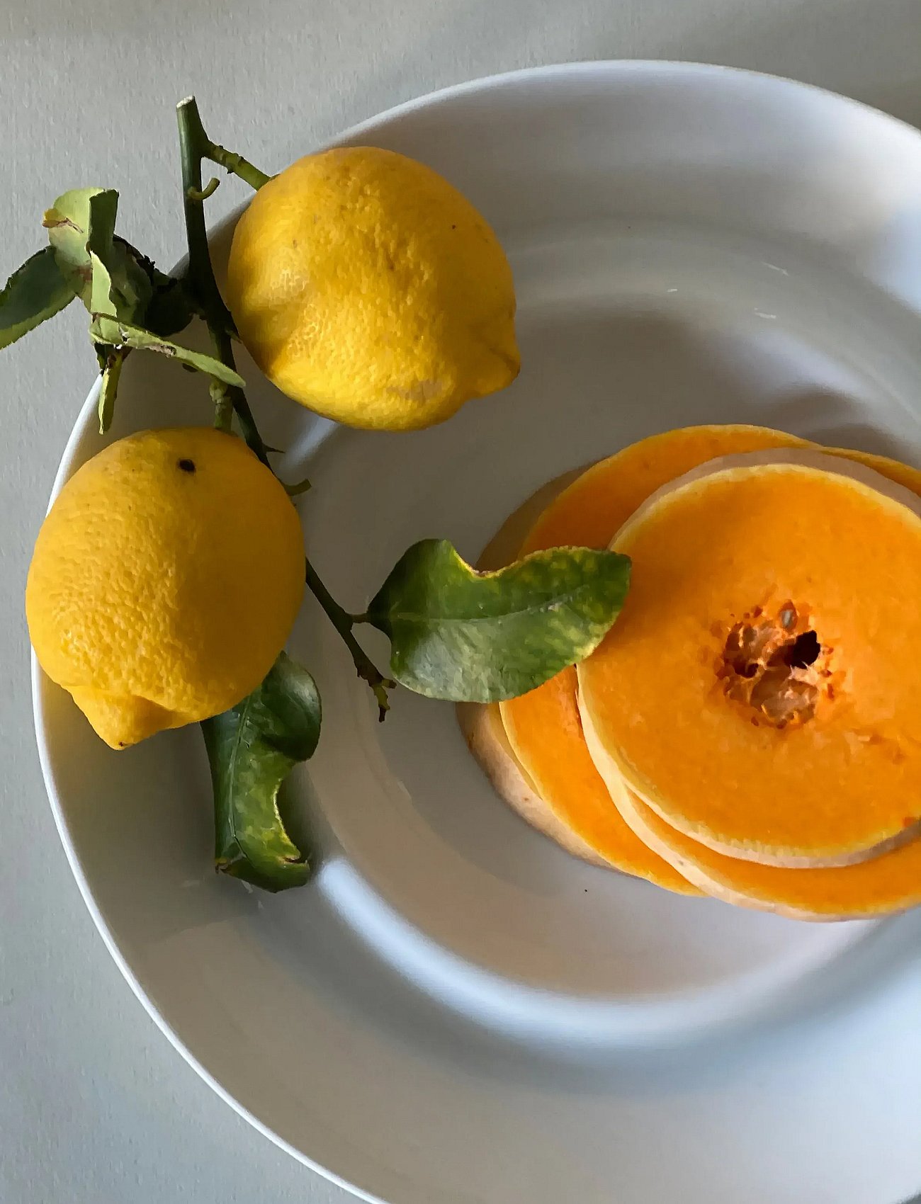 To citroner på en gren med blade og runde skiver af orange græskar på et hvidt serveringsfad.