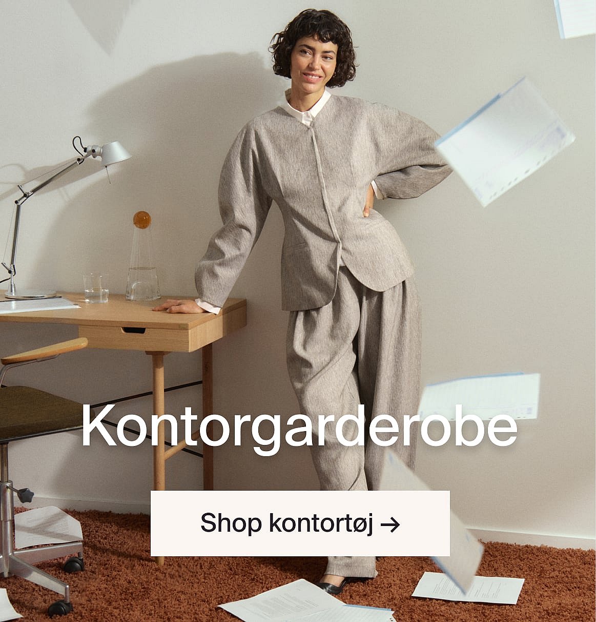 En smilende kvinde i et gråt jakkesæt læner sig op ad et skrivebord på et kontor. Tekst: Kontorgarderobe.