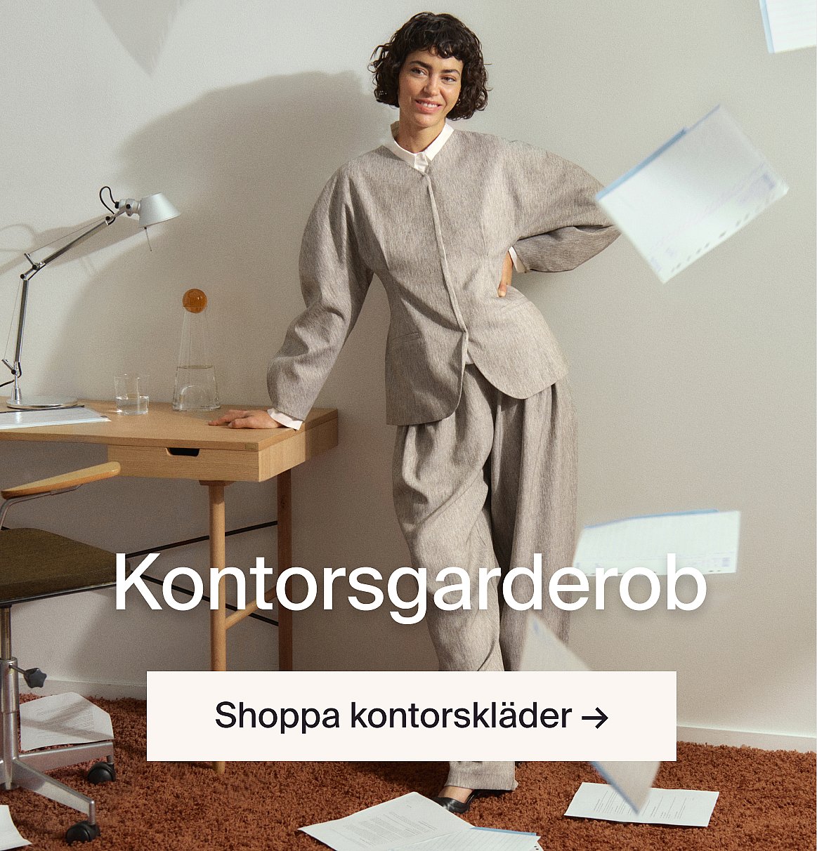 En leende kvinna i grå kostym lutar sig mot ett skrivbord på ett kontor. Text: Kontorsgarderob.