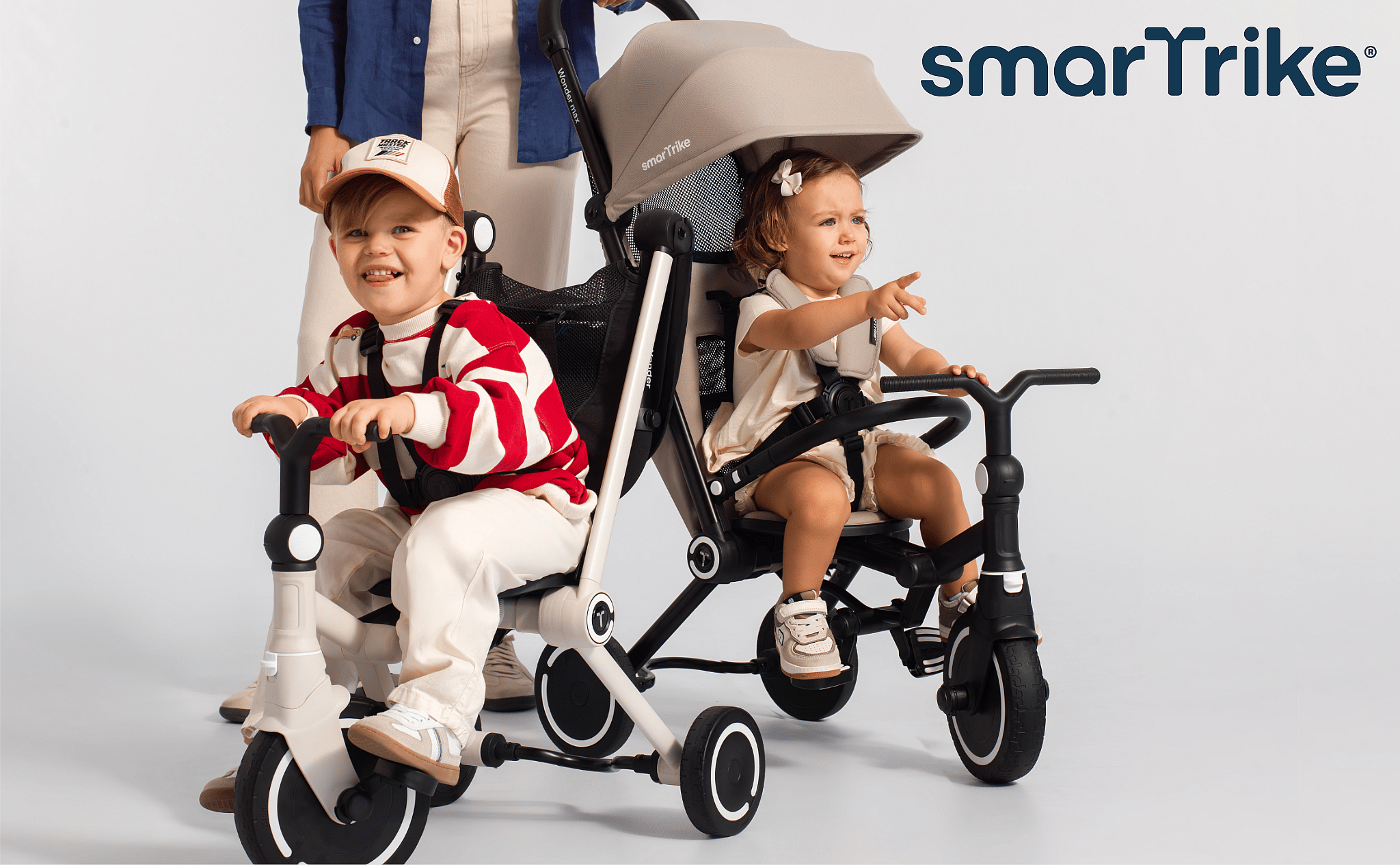 Two toddlers on smart trikes, one smiling, one pointing, with an adult behind them in a studio setting.