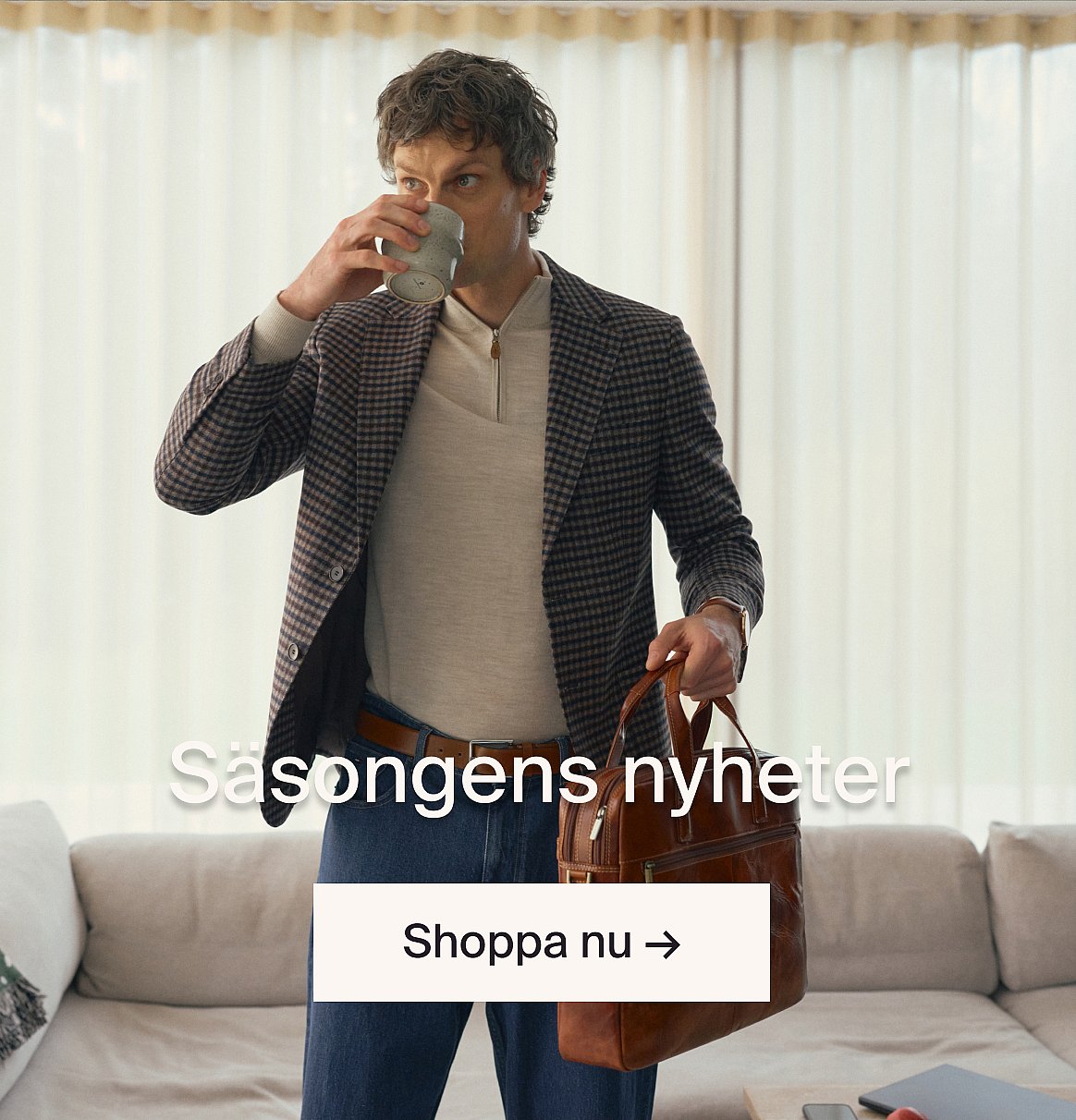 Person i vit kortärmad broderie anglaise-skjorta och blå jeans, händerna i fickorna, står mot en ljus trävägg.