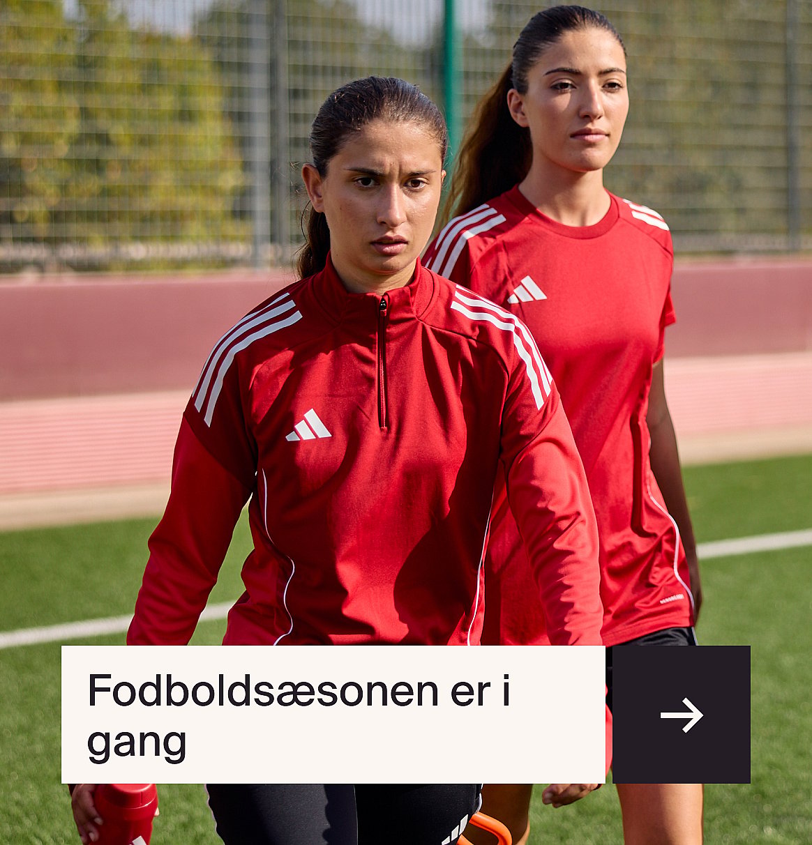 To kvinder i rødt adidas-sportstøj på en fodboldbane. Den ene har en langærmet trøje på, den anden en T-shirt.