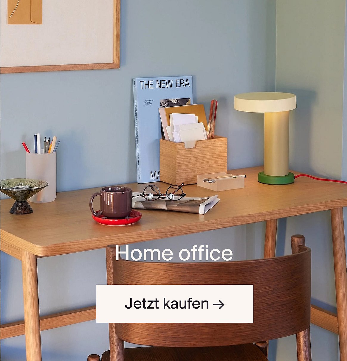 Ein Homeoffice-Schreibtisch aus hellem Holz mit einer Lampe, einer Tasse, Gläsern und Büromaterial vor einer blauen Wand. Text: Homeoffice, Jetzt kaufen.