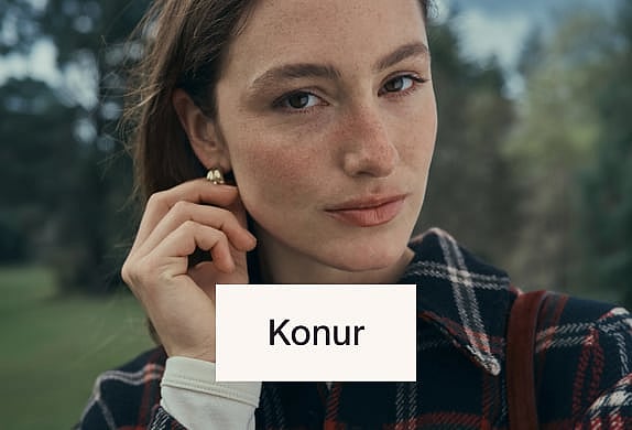 Kona í rauðri ofinni húfu og sólgleraugum, horfir upp á sólskinsströnd með hafi og klettum.
