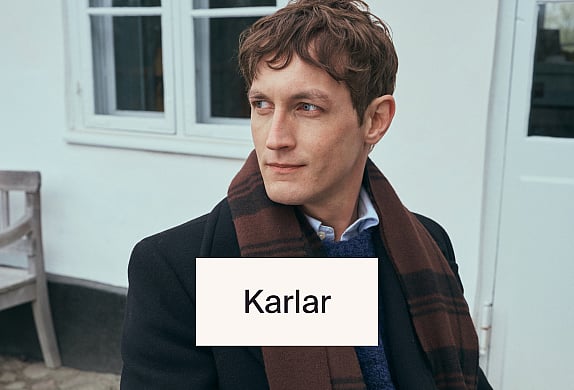 Maður í sólgleraugum, röndóttum bol og hvítum buxum, með flösku í hendi, situr í rauðum stól við sjóinn.