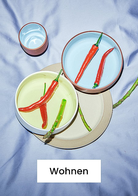 Hellblaue und gelbe Schüsseln mit roten Chilischoten und Spargel, eine Tasse und ein beiger Teller auf einem hellblauen Tuch. Text: Zuhause