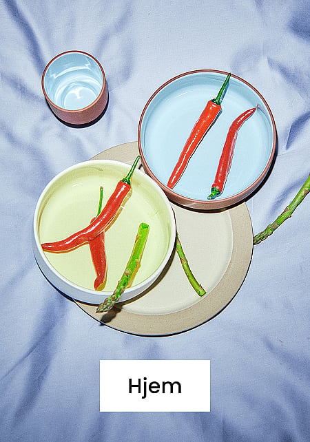 Lyseblå og gule skåler med røde chilier og asparges, en kopp og en beige tallerken på et lyseblått stoff. Tekst: Hjem