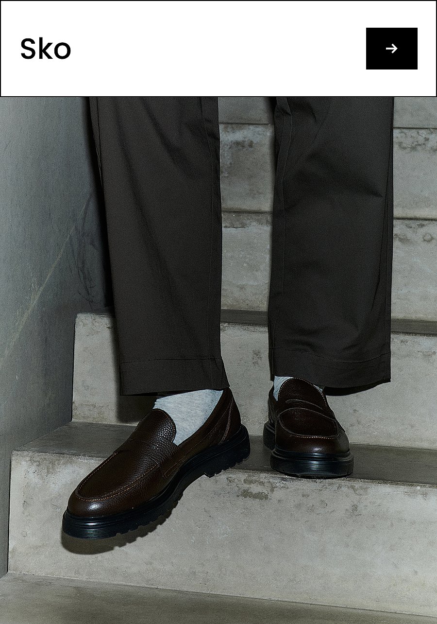 En persons ben i mørke bukser og lyse sokker, iført brune skinnloafers med tykke svarte såler, stående på betong. Tekst: Sko