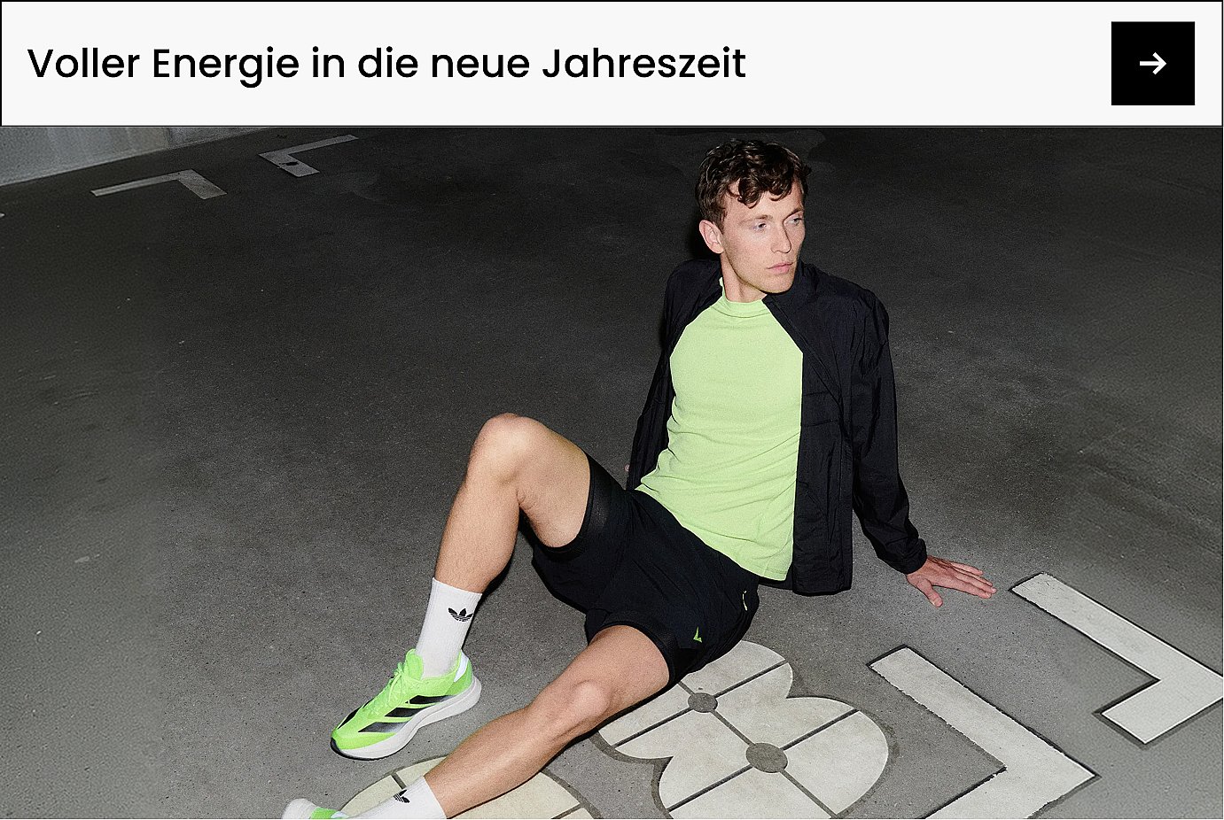 Ein Mann in neongrünem Sport-Top und -Schuhen, schwarzer Jacke und Shorts, der auf einer dunklen Betonfläche mit weißen Markierungen sitzt. Sport.